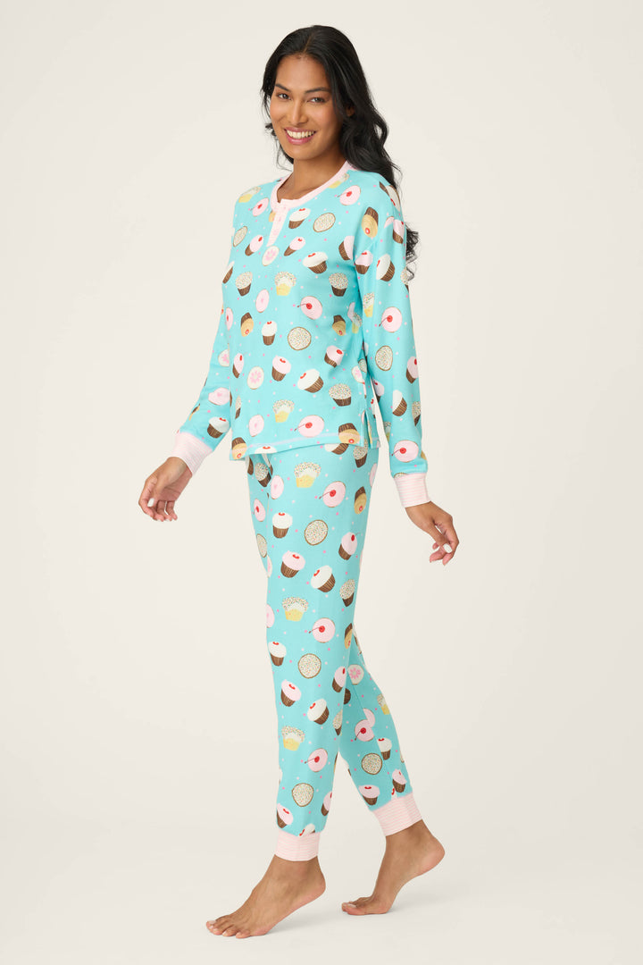 A smiling woman with long dark hair stands barefoot in a P.J. Salvage Sweet Life Cupcake Print PJ Set, featuring a colorful donut and cupcake pattern. She poses against a plain, light-colored background.