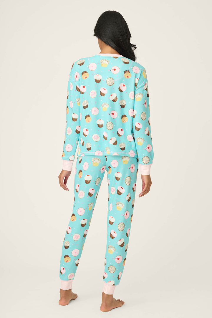 A person stands barefoot facing away, wearing the P.J. Salvage Sweet Life Cupcake Print PJ Set—light blue pajamas with a colorful cupcake and donut pattern, finished with light pink cuffs at the wrists and ankles.