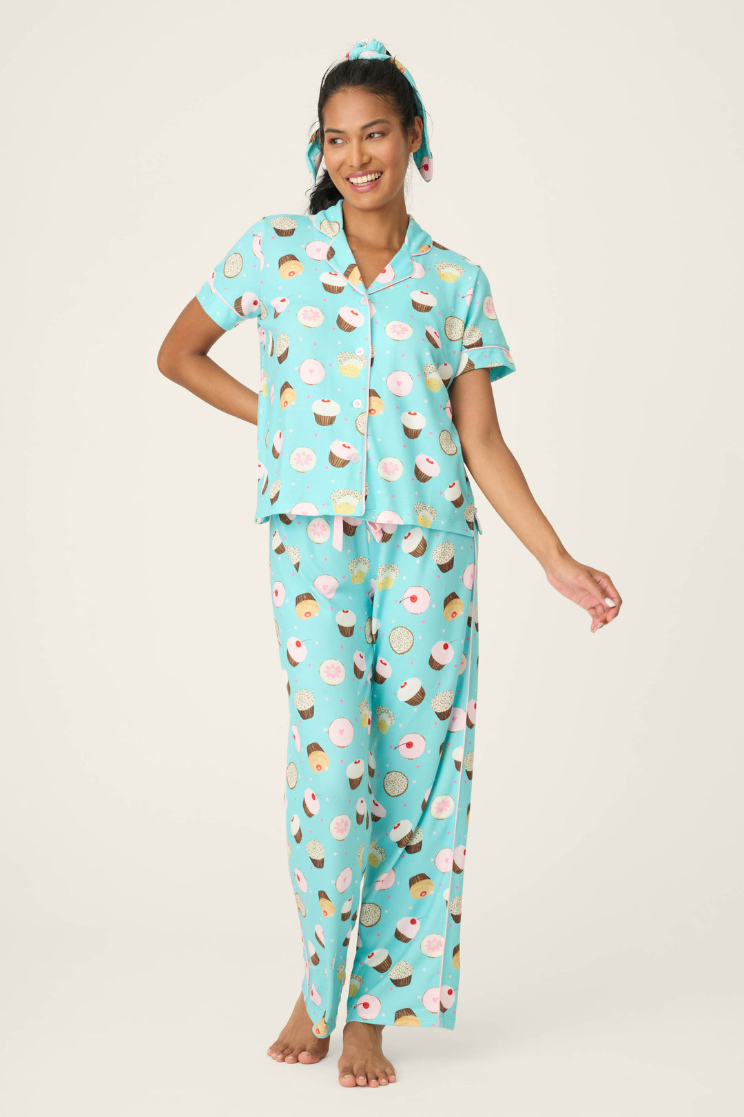 A woman smiles barefoot in the P.J. Salvage Sweet Life Cupcake Print 3-Piece PJ Set, featuring a light blue short-sleeve button-up top, long pants with cupcake and donut prints, and a matching scrunchie in her hair.