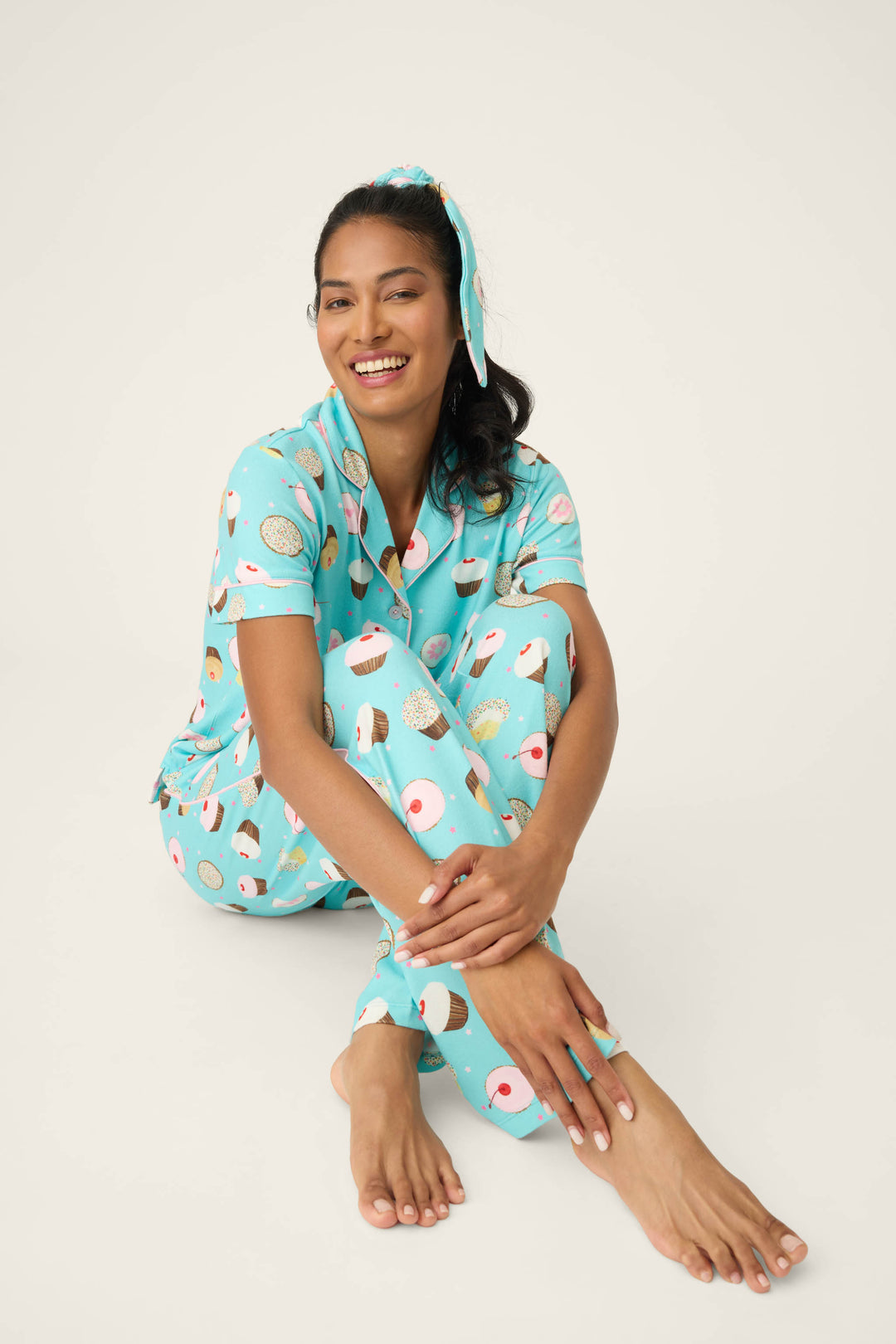 A woman sits on the floor, smiling with her arms resting on her knees. She wears the P.J. Salvage Sweet Life Cupcake Print 3-Piece PJ Set with a matching headband against a plain, light-colored background.