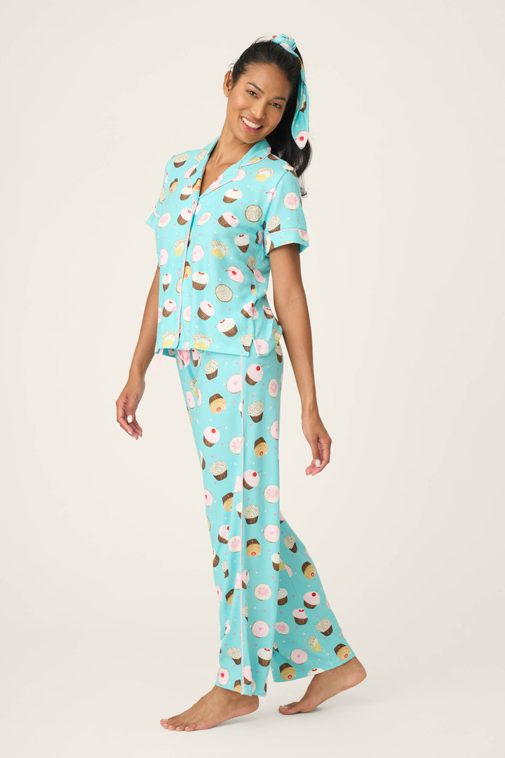 A woman smiles barefoot in the P.J. Salvage Sweet Life Cupcake Print 3-Piece PJ Set, featuring light blue pajama pants, a matching short-sleeve top with a cupcake print, and a coordinating hair scrunchie, against a plain white background.
