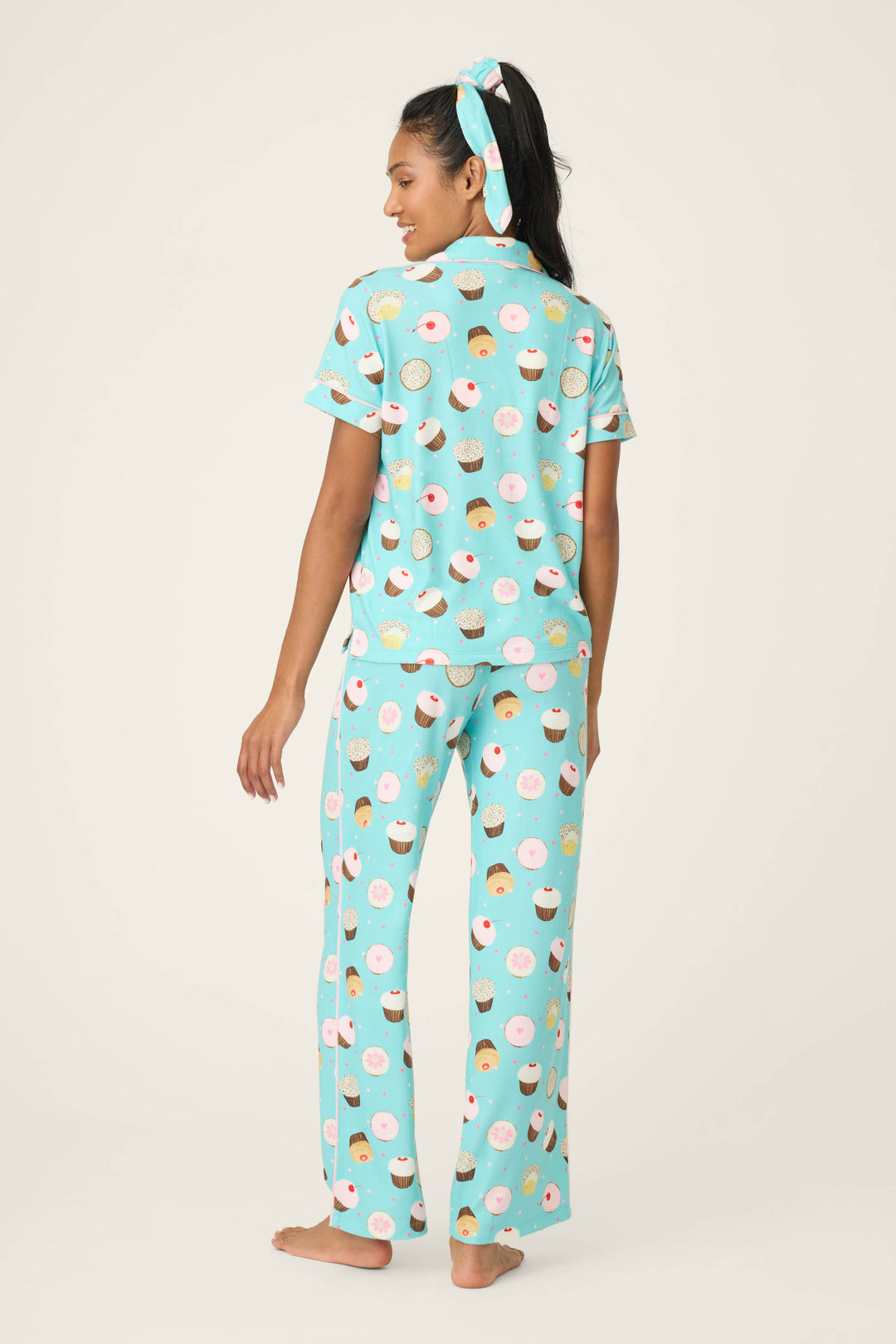 A person is barefoot, facing away, wearing the P.J. Salvage Sweet Life Cupcake Print 3-Piece PJ Set with light blue matching cupcake-print pajamas and a matching hair tie. The sleep set includes a short-sleeve top and long pants. Plain light background.