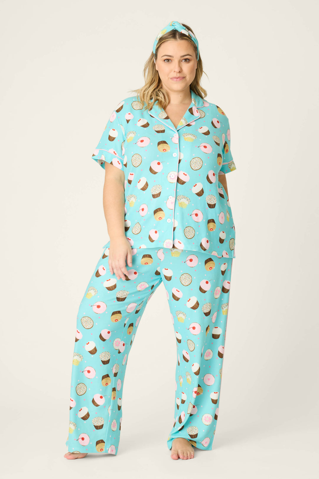 A woman stands barefoot in the P.J. Salvage Sweet Life Cupcake Print 3-Piece Plus Size PJ Set—light blue with donut and cupcake print, matching pants, shirt, and headband—against a plain light background.