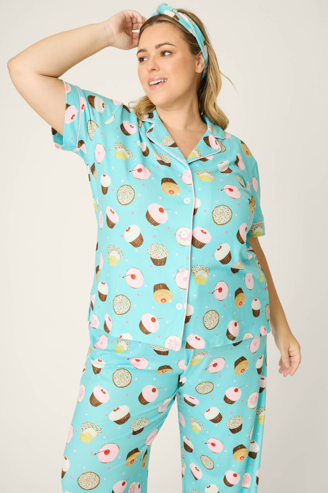 A woman in the P.J. Salvage Sweet Life Cupcake Print 3-Piece Plus Size PJ Set, featuring turquoise fabric with colorful donut and cupcake designs and a matching headband, stands smiling with one arm raised, looking to the side.