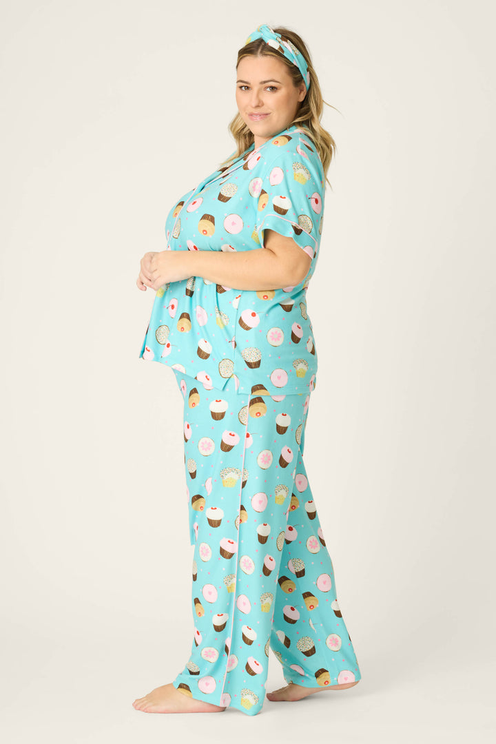A person stands barefoot, sideways, looking at the camera in the P.J. Salvage Sweet Life Cupcake Print 3-Piece Plus Size PJ Set, featuring a matching loose-fit blue short-sleeve top, pants, and a coordinating headband with a dessert print.