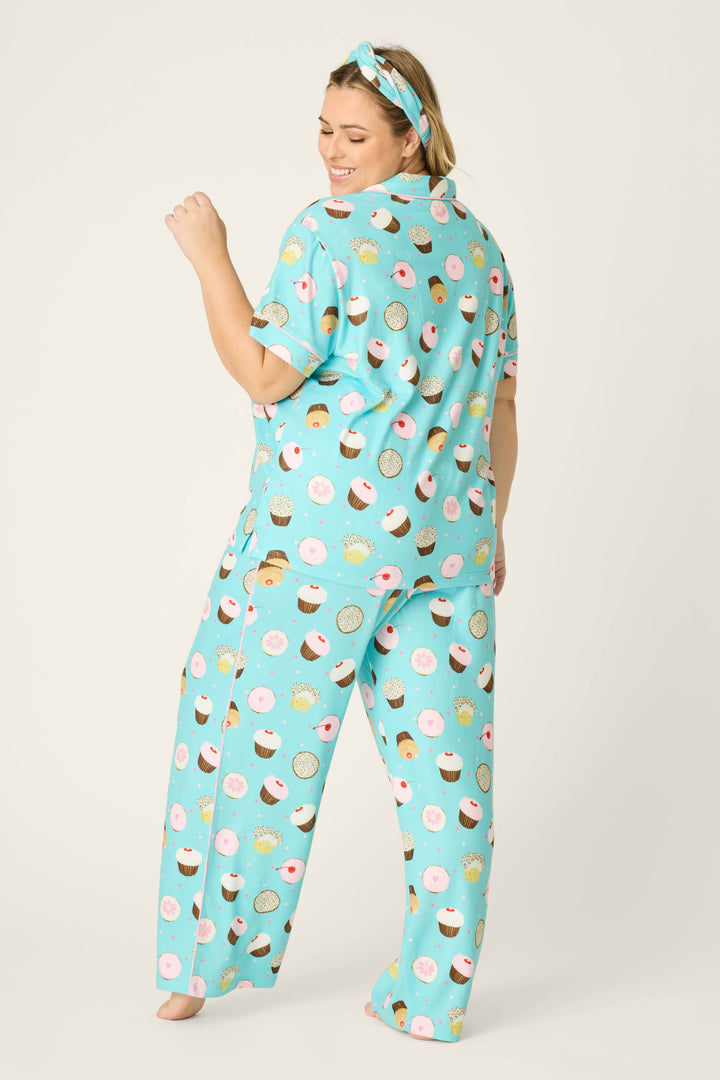 A woman stands barefoot with her back to the camera, wearing the P.J. Salvage Sweet Life Cupcake Print 3-Piece Plus Size PJ Set and a matching headband with a colorful cupcake and donut print, smiling over her shoulder against a plain white background.