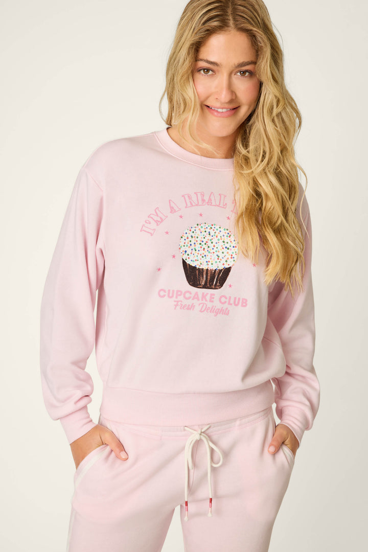 A woman with long blonde hair wears the P.J. Salvage Sweet Life "I'm A Real Treat" Lounge Set in light pink, featuring a cupcake graphic and playful text on the sweatshirt.