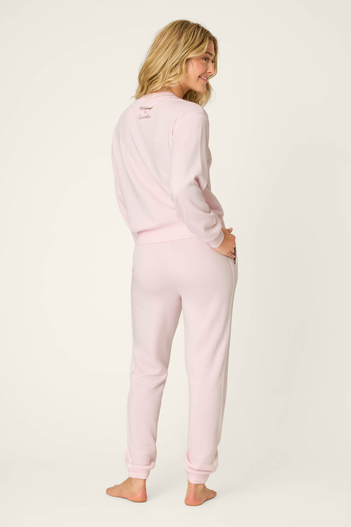 A woman stands barefoot with one hand on her hip, smiling over her shoulder while wearing the P.J. Salvage Sweet Life "I'm A Real Treat" Lounge Set in light pink against an off-white background.