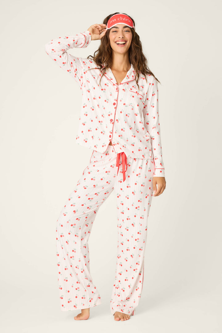 A person stands barefoot and smiling, wearing the P.J. Salvage Valentine's Day Cherry Print 3-Piece PJ Set with red trim and a matching red sleep mask on their forehead, posing with one hand touching the mask.