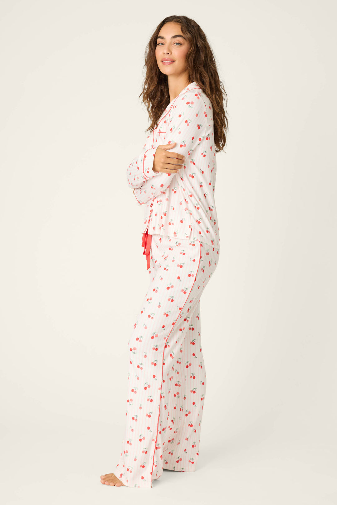 A woman with long wavy hair poses barefoot in a side view, wearing the P.J. Salvage Valentine's Day Cherry Print 3-Piece PJ Set—long-sleeve top and matching pants—against a plain background, looking relaxed.