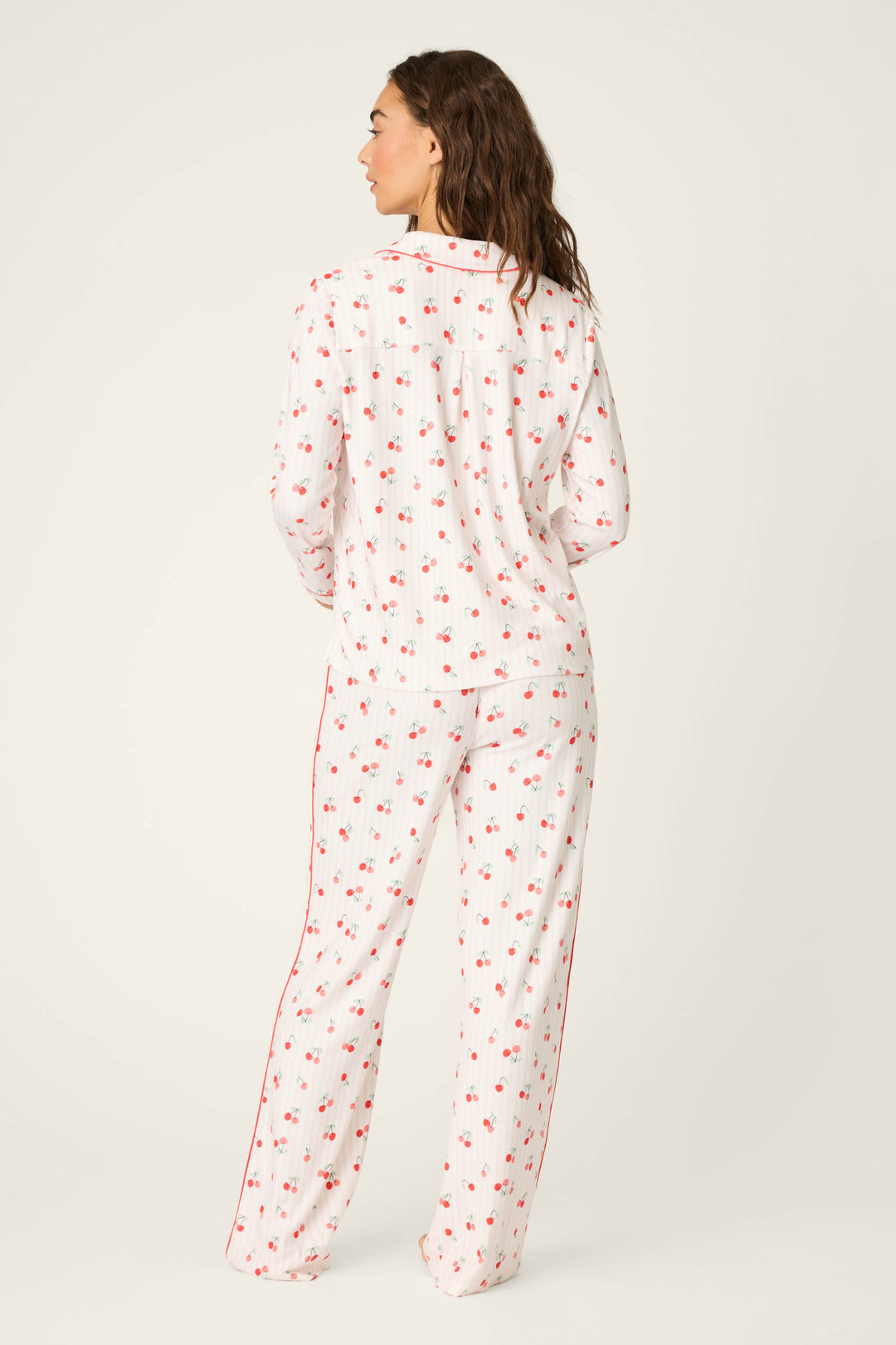 A person stands with their back to the camera, wearing the P.J. Salvage Valentine's Day Cherry Print 3-Piece PJ Set featuring a white base, red cherry prints, and red piping, set against a plain light background.