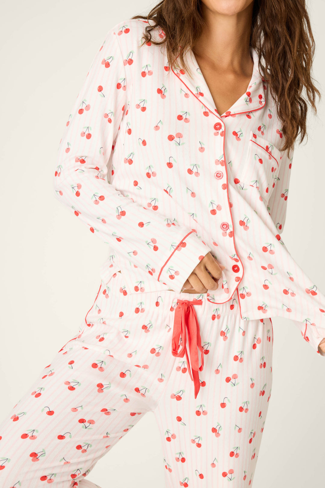 A woman models the P.J. Salvage Valentine's Day Cherry Print 3-Piece PJ Set, featuring white fabric with red cherries, red piping, red buttons, and a red waist drawstring; she stands angled with one leg raised.