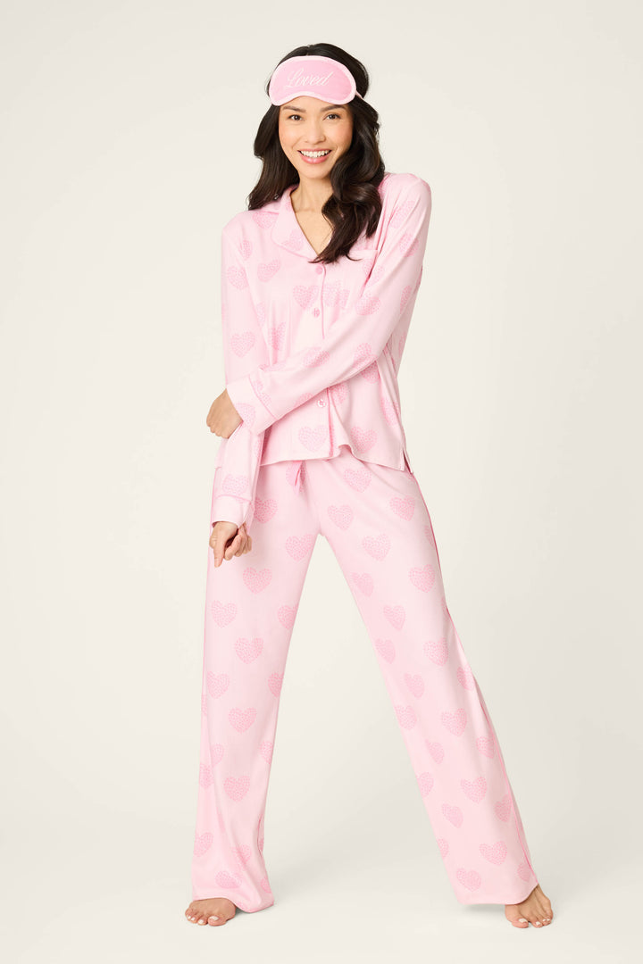 A woman stands barefoot, smiling in the P.J. Salvage Valentine's Day Leopard Hearts Print 3-Piece PJ Set with a matching pink sleep mask. The background is plain and light-colored.