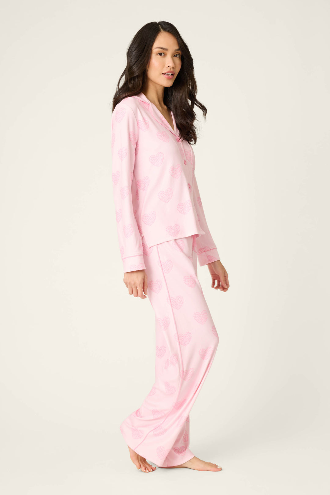 A woman with long, dark hair stands barefoot on a plain background, wearing the P.J. Salvage Valentine's Day Leopard Hearts Print 3-Piece PJ Set featuring a light pink button-up top and matching long pants. She is facing slightly to the side.