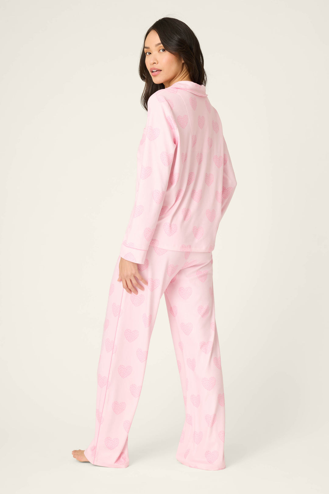 A woman stands barefoot, facing away from the camera, wearing P.J. Salvage's Valentine's Day Leopard Hearts Print 3-Piece PJ Set in light pink. She glances over her shoulder against an off-white background.