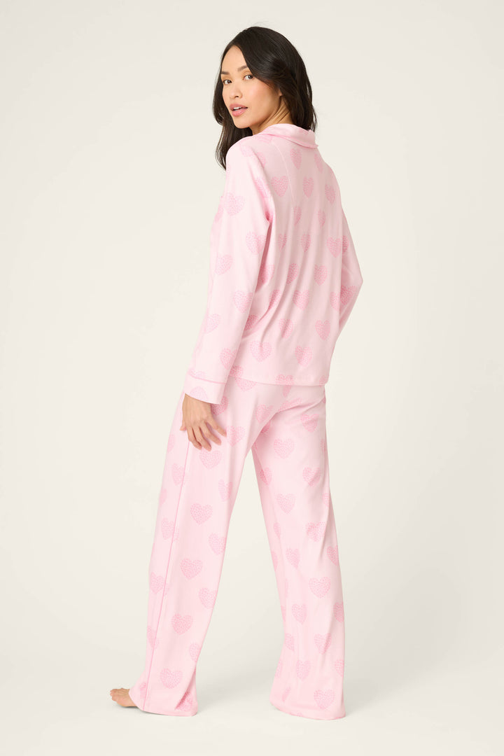 A woman stands barefoot, facing away from the camera, wearing P.J. Salvage's Valentine's Day Leopard Hearts Print 3-Piece PJ Set in light pink. She glances over her shoulder against an off-white background.