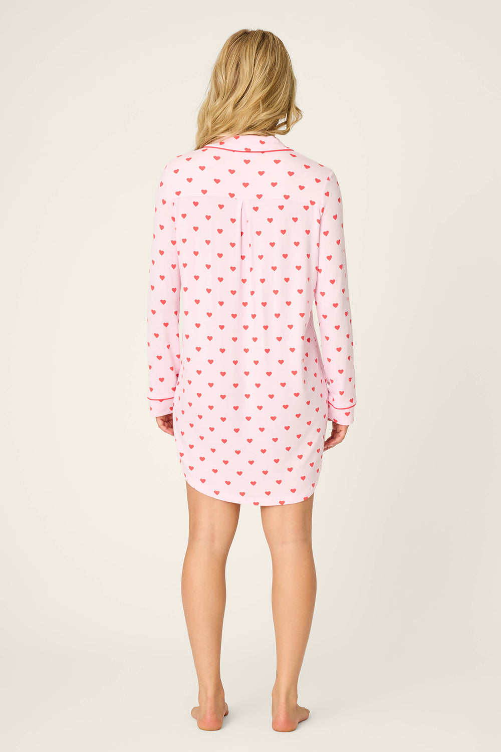 A woman stands barefoot facing away, wearing the P.J. Salvage Wildest Dreams Heart Print Nightshirt—a long-sleeved, knee-length nightshirt with a white base and red hearts. Her hair is loose against a plain, light background.