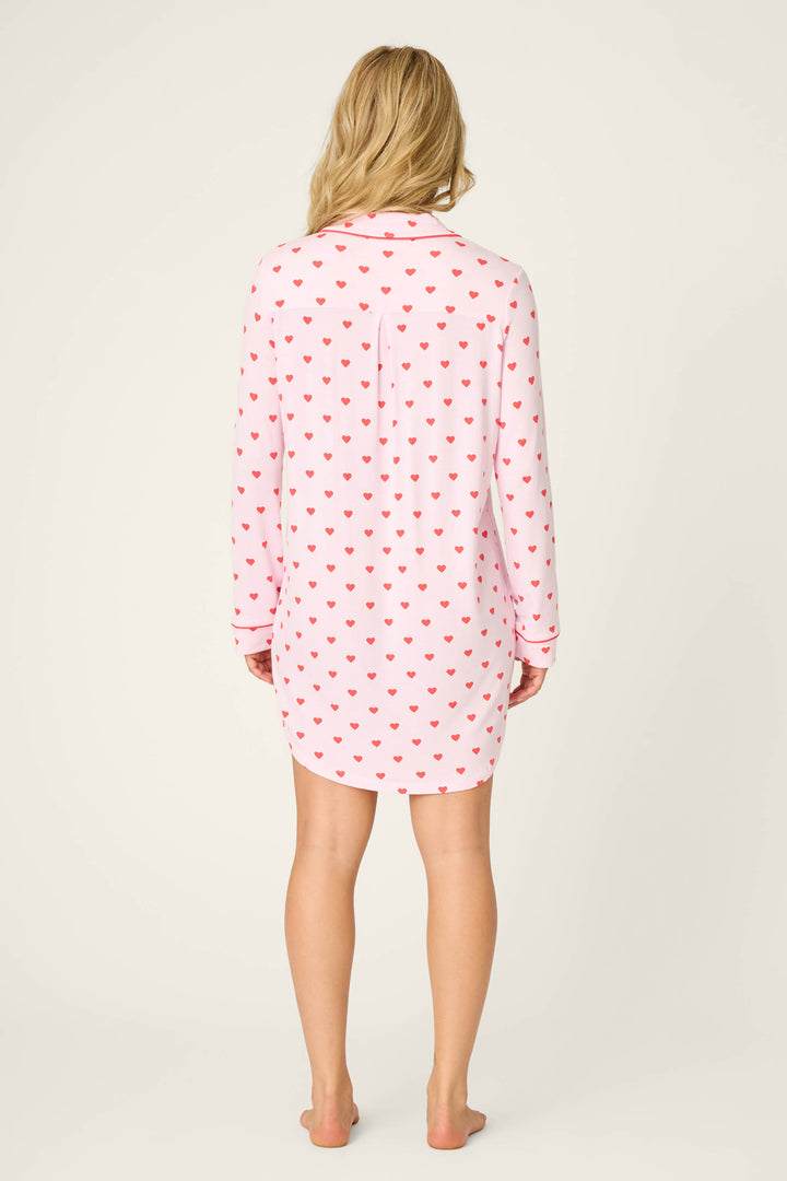 A woman stands barefoot facing away, wearing the P.J. Salvage Wildest Dreams Heart Print Nightshirt—a long-sleeved, knee-length nightshirt with a white base and red hearts. Her hair is loose against a plain, light background.