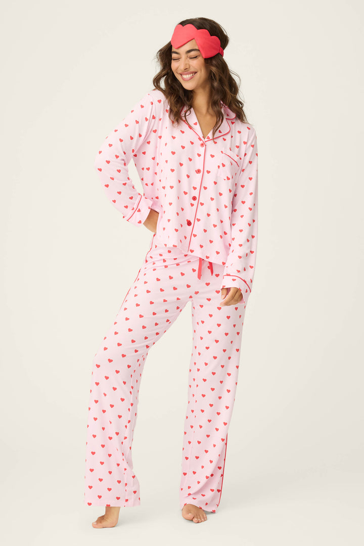 A person stands barefoot in the P.J. Salvage Wildest Dreams Heart Print 3-Piece PJ Set, featuring pink pajamas with red hearts, button-up top with piping, and a matching sleep mask. The background is plain and light-colored.