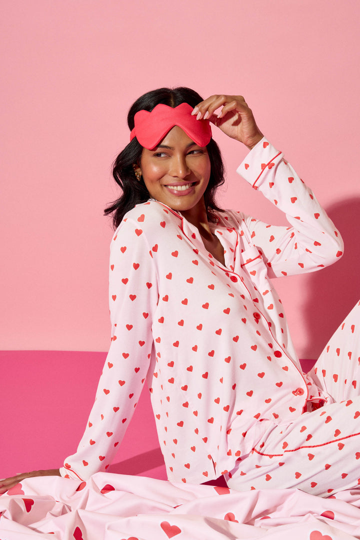 A person smiles at the camera, sitting on a pink surface against a pink background, wearing the P.J. Salvage Wildest Dreams Heart Print 3-Piece PJ Set and holding a red heart-shaped sleep mask above their head.