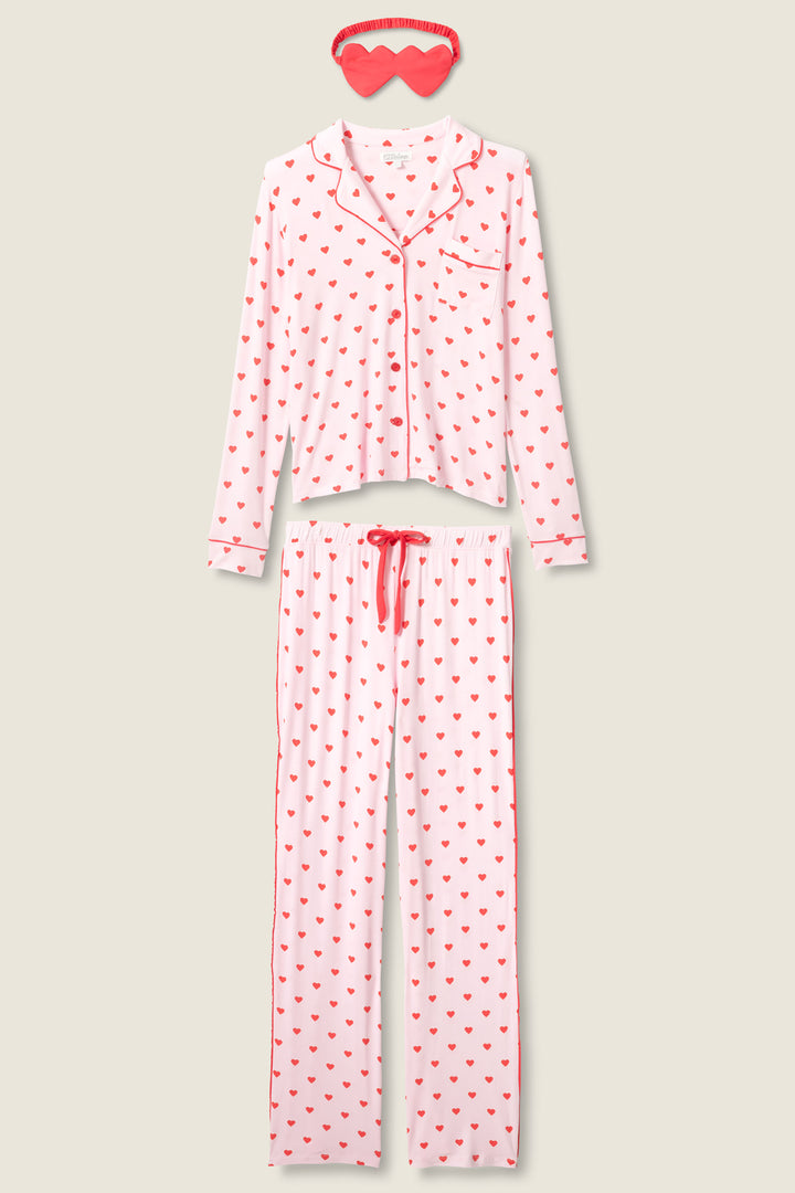 The P.J. Salvage Wildest Dreams Heart Print 3-Piece PJ Set features a pink long-sleeve button-up top, drawstring pants with red heart prints, and a matching sleep mask, displayed on a light beige background.