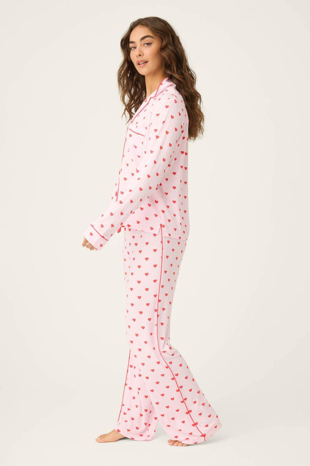 A woman stands barefoot in the P.J. Salvage Wildest Dreams Heart Print 3-Piece PJ Set, featuring a matching light pink long-sleeve button-up top and full-length pants with red heart patterns. She faces sideways against a plain background.