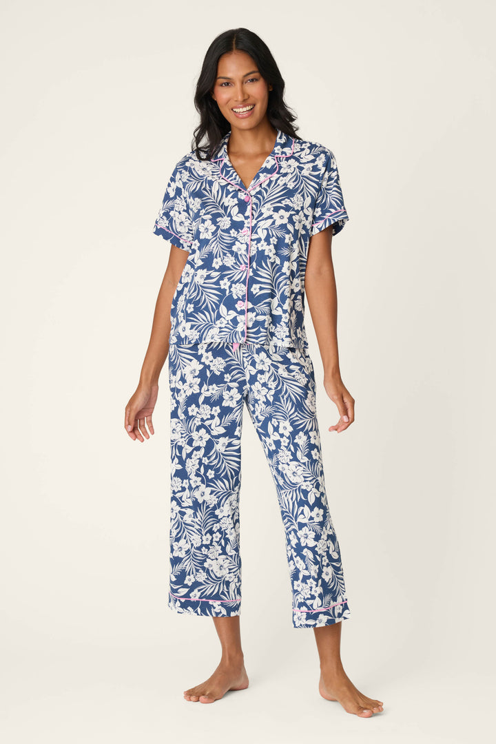 A woman stands barefoot, smiling, in the P.J. Salvage Cotton Jersey Midnight Garden Cropped PJ Set—featuring a matching blue and white floral design with short sleeves and cropped pants—against a plain light background.