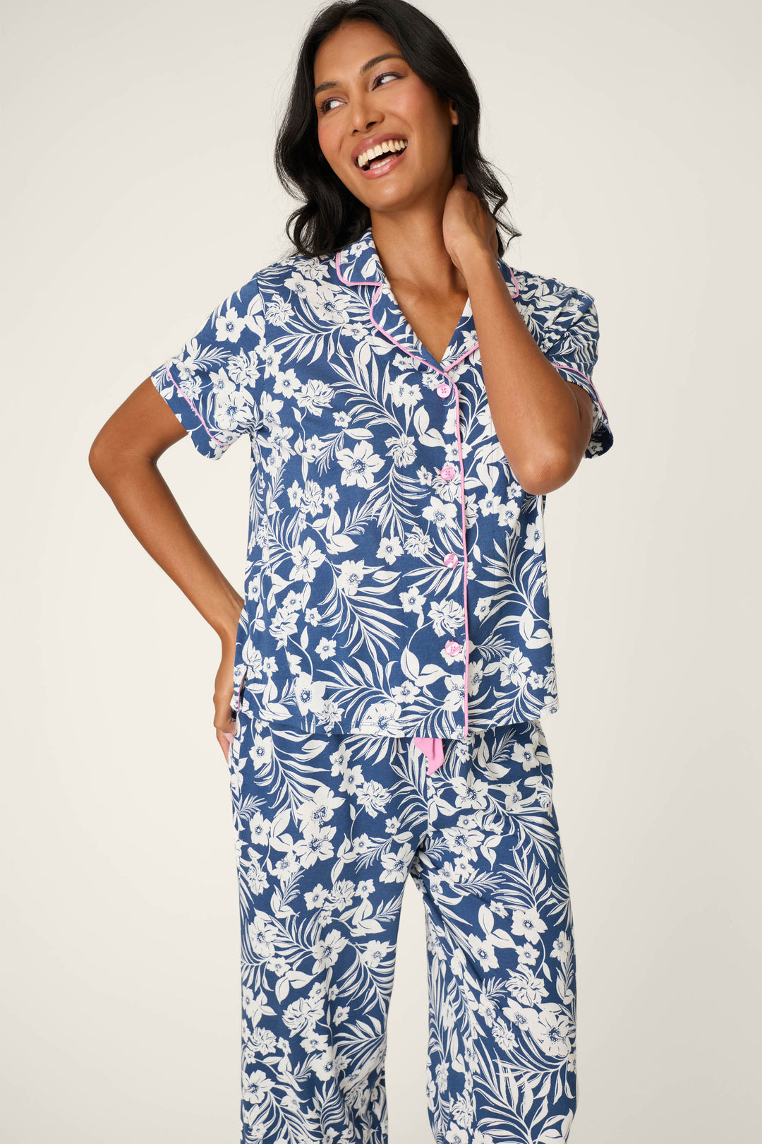 A woman smiles in the P.J. Salvage Cotton Jersey Midnight Garden Cropped PJ Set, featuring blue and white florals with short sleeves and pink piping, standing against a plain light background.