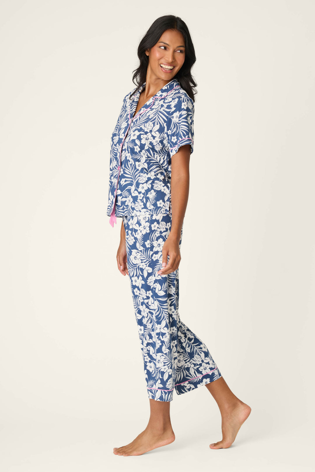 A woman stands barefoot, smiling and looking to the side, wearing the P.J. Salvage Cotton Jersey Midnight Garden Cropped PJ Set—a blue pajama set with a white floral pattern, short sleeves, and cropped pants—against a plain light background.