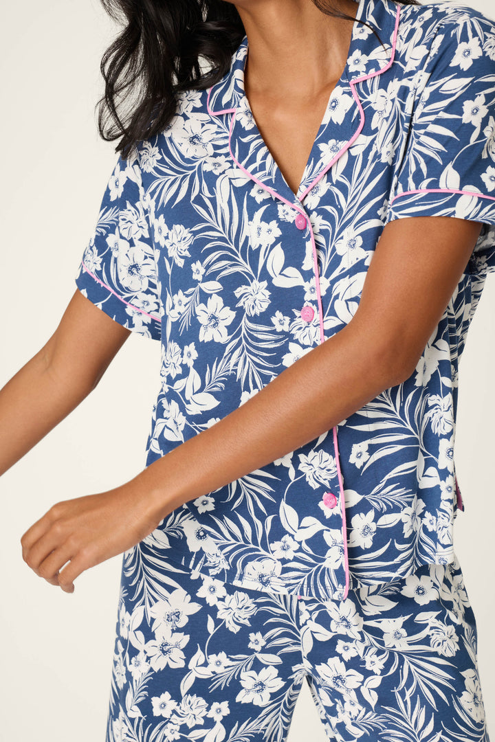 A person, shown from the shoulders down with arms slightly bent and moving, wears the P.J. Salvage Cotton Jersey Midnight Garden Cropped PJ Set—blue, short-sleeved, with a white floral print and pink piping.