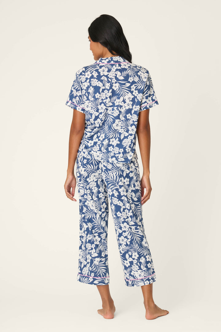 A woman with long dark hair, barefoot and facing away, wears the P.J. Salvage Cotton Jersey Midnight Garden Cropped PJ Set—short sleeves and cropped pants in a blue and white floral print—against a plain light background.