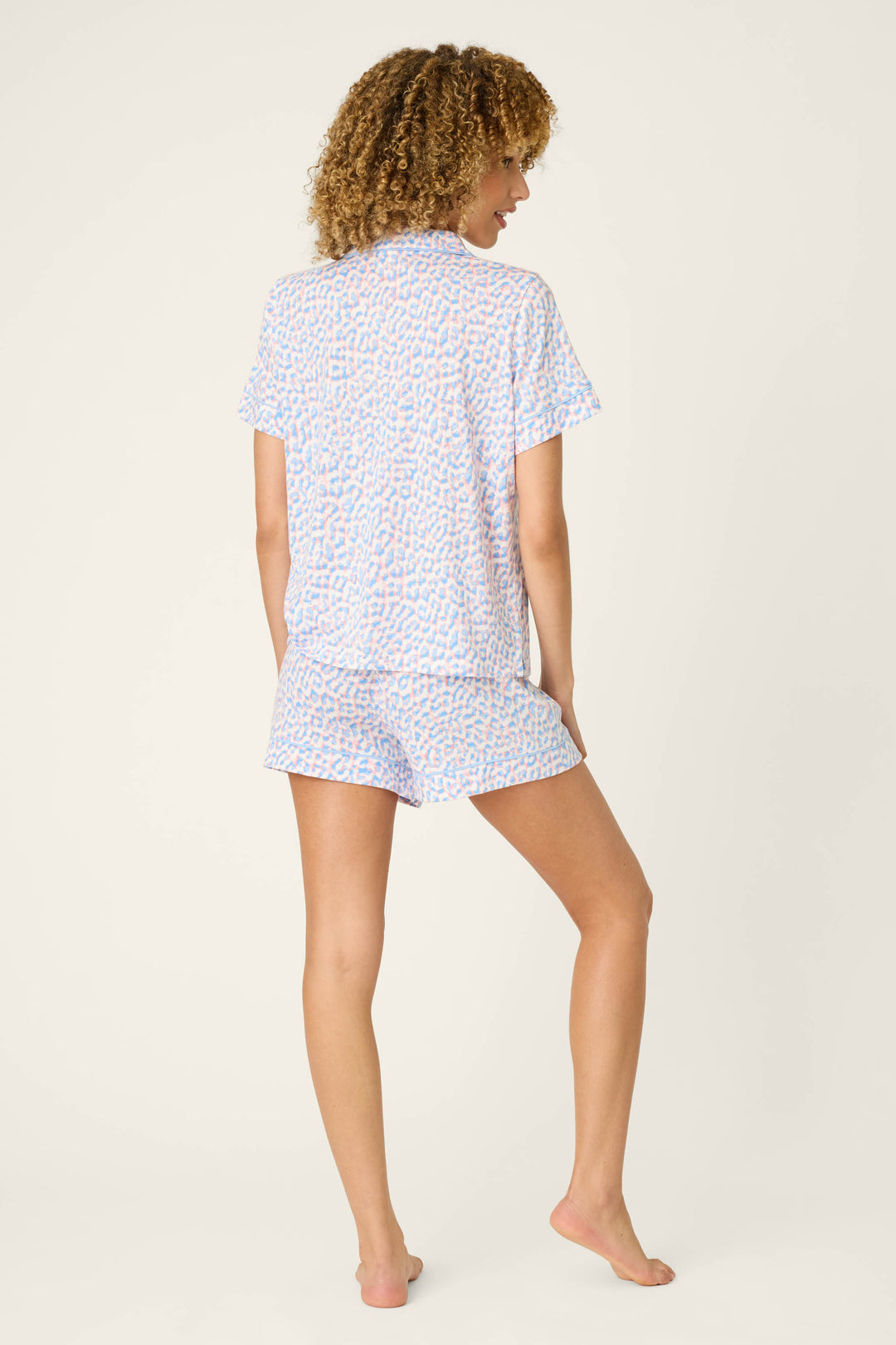 A person stands barefoot facing away, wearing the P.J. Salvage Animal Lover Leopard Print 3-Piece Shorts Set with a light animal print pattern. The background is plain and light-colored.