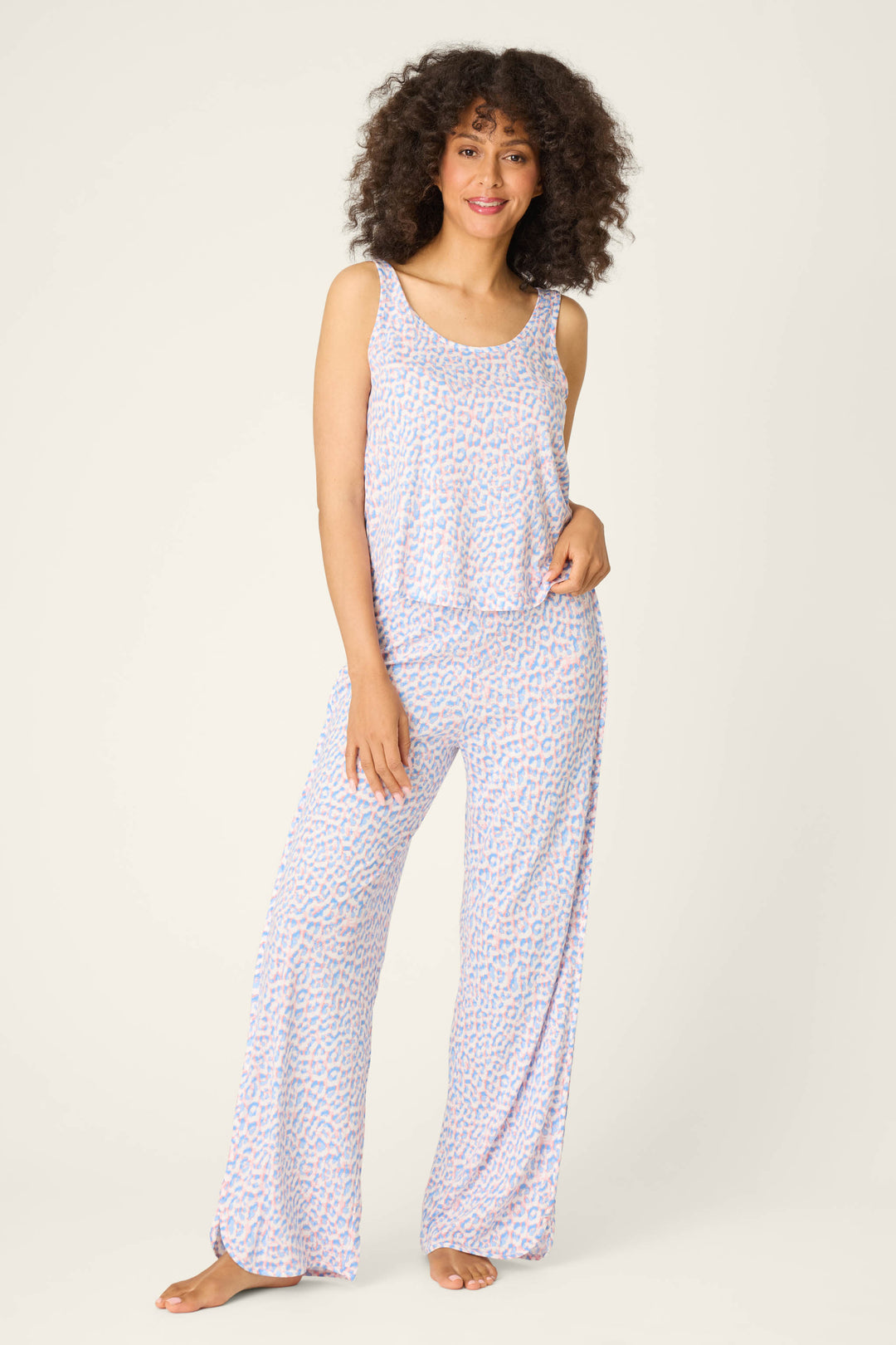 A woman with curly hair, barefoot against a plain backdrop, smiles with one hand on her hip while wearing P.J. Salvage Animal Lover Leopard Print Open Leg Pants and a loose, sleeveless top.