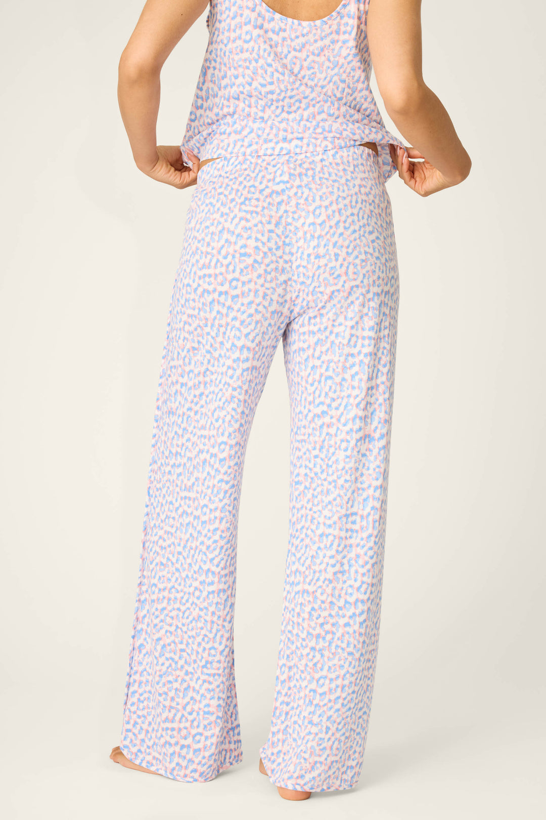 A person stands barefoot with their back to the camera, wearing P.J. Salvage’s Animal Lover Leopard Print Open Leg Pants in a light purple and pink leopard pattern, adjusting the waistband.