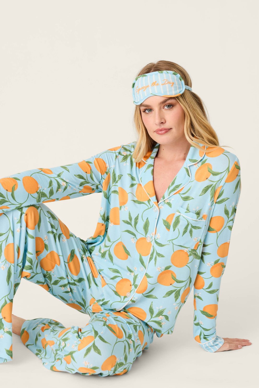 A woman with long blonde hair sits on the floor, wearing the P.J. Salvage Citrus Dreams Morning Squeeze Long 3-Piece PJ Set—stylish blue citrus-print sleepwear with a matching sleep mask—against a plain light background.