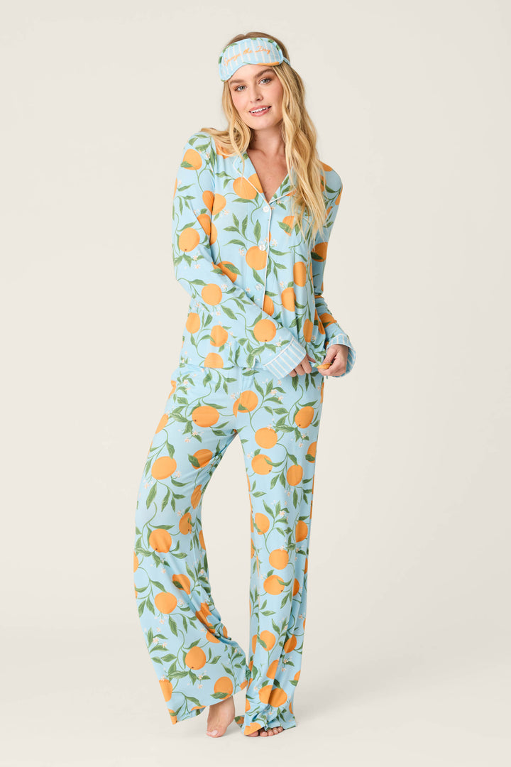A person stands barefoot in the Citrus Dreams Morning Squeeze Long 3-Piece PJ Set by P.J. Salvage, featuring a playful citrus print and matching sleep mask, against a plain light background—showcasing stylish women's sleepwear.