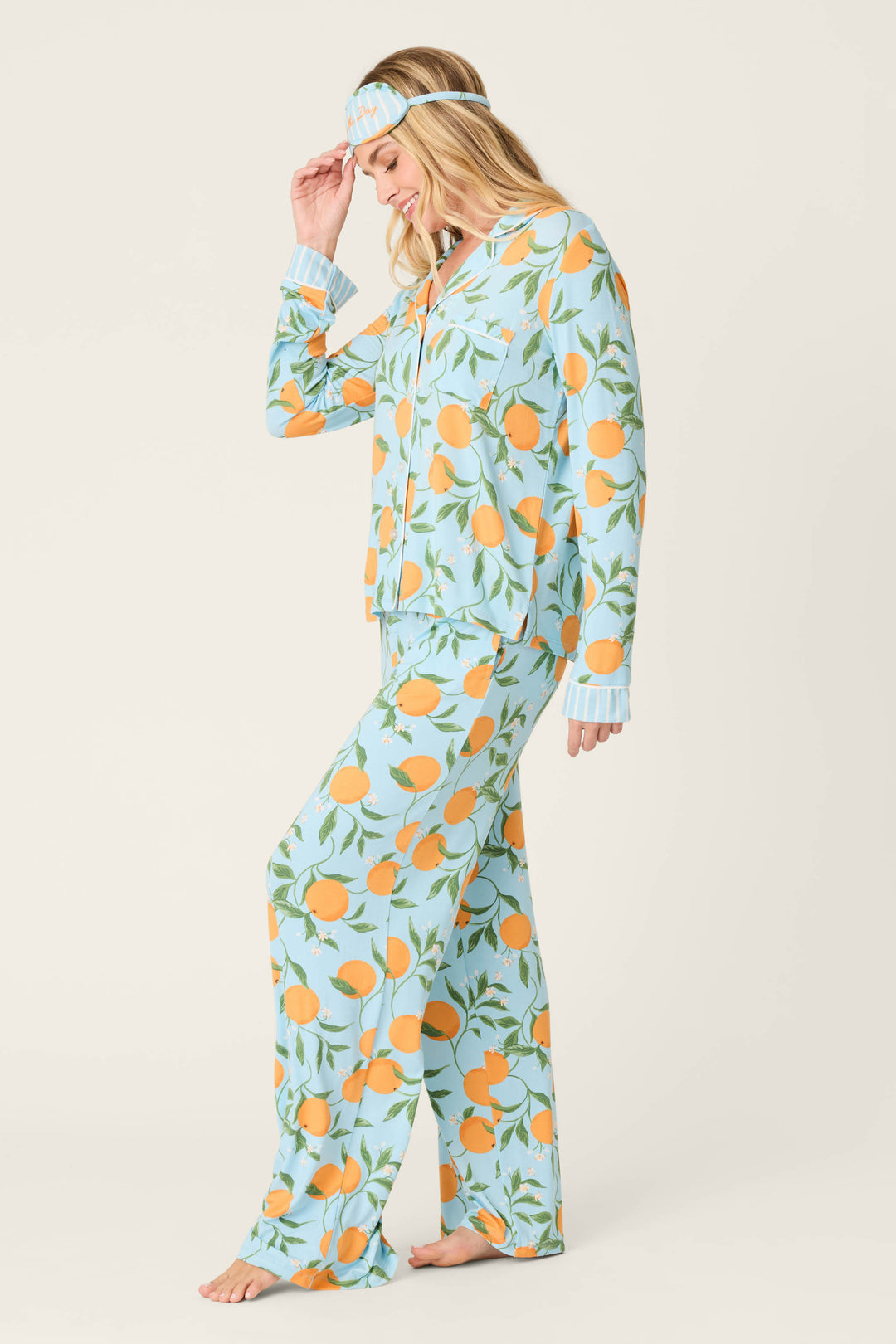 A woman smiles barefoot in the P.J. Salvage Citrus Dreams Morning Squeeze Long 3-Piece PJ Set, featuring a light blue citrus print and matching sleep mask. She gently holds the mask, standing against an off-white background that highlights stylish women's sleepwear.