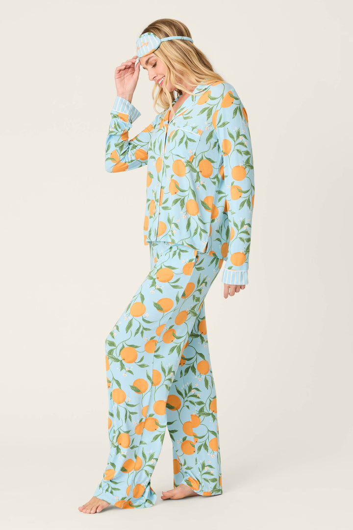 A woman smiles barefoot in the P.J. Salvage Citrus Dreams Morning Squeeze Long 3-Piece PJ Set, featuring a light blue citrus print and matching sleep mask. She gently holds the mask, standing against an off-white background that highlights stylish women's sleepwear.