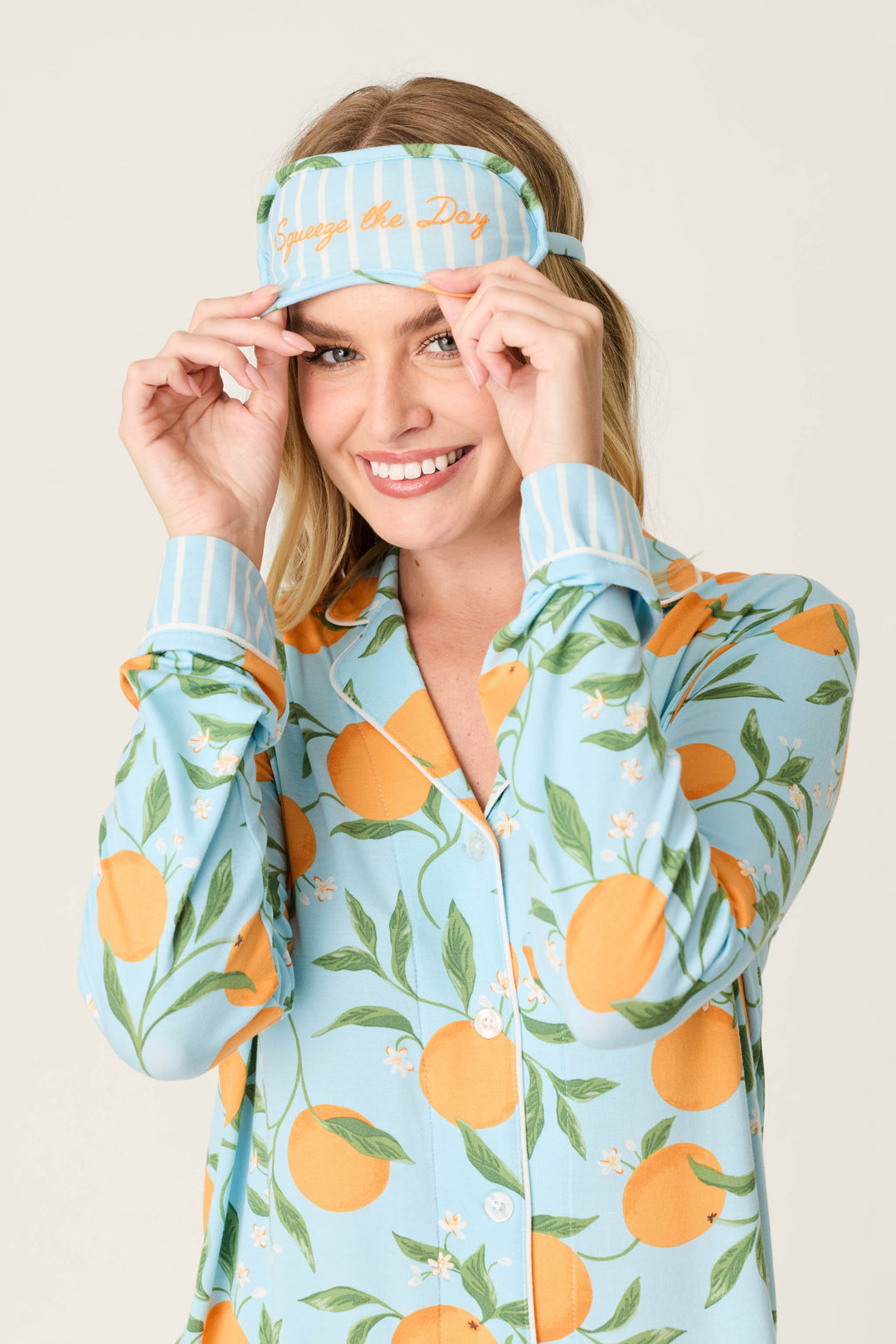 A person in the Citrus Dreams Morning Squeeze Long 3-Piece PJ Set by P.J. Salvage smiles while adjusting a striped sleep mask that says Squeeze the Day, showing off their love for playful, stylish women’s sleepwear.