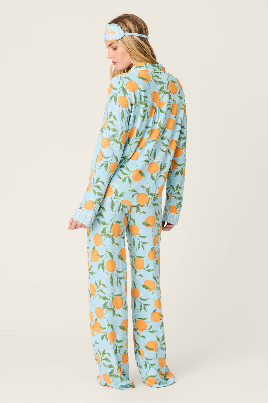 A woman stands turned slightly away, wearing the Citrus Dreams Morning Squeeze Long 3-Piece PJ Set by P.J. Salvage, featuring a blue top and citrus-print sleep mask with oranges and green leaves against a plain, light backdrop.
