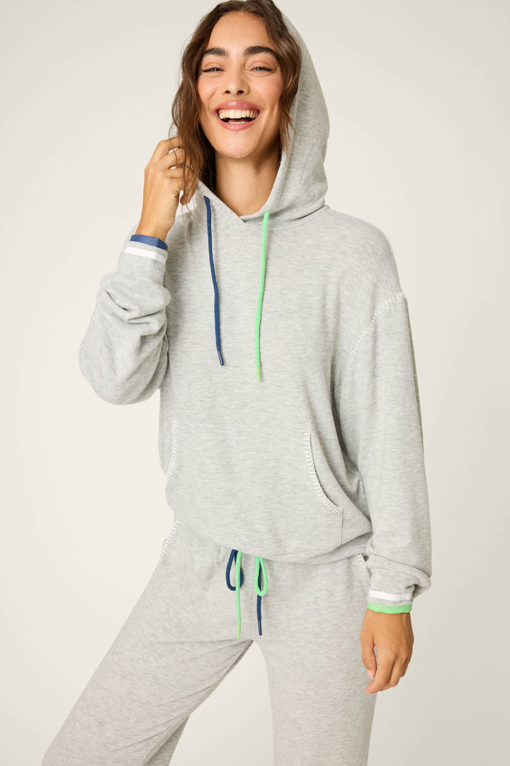 A person wears the P.J. Salvage C’est La Vie Heather Grey Hoodie with green and blue drawstrings, paired with matching sweatpants featuring a green drawstring, smiling and touching their hair.