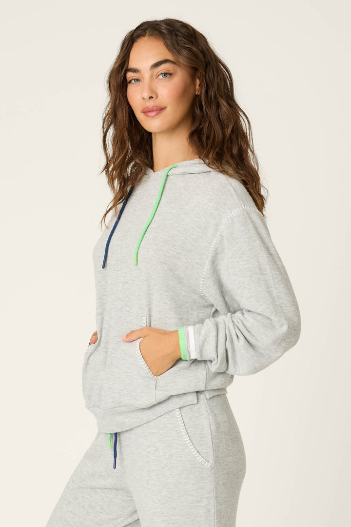 A person with long wavy brown hair wears the P.J. Salvage C'est La Vie Heather Grey Hoodie, featuring blue and green drawstrings, green and white cuff stripes, and matching sweatpants, with hands in the pockets.