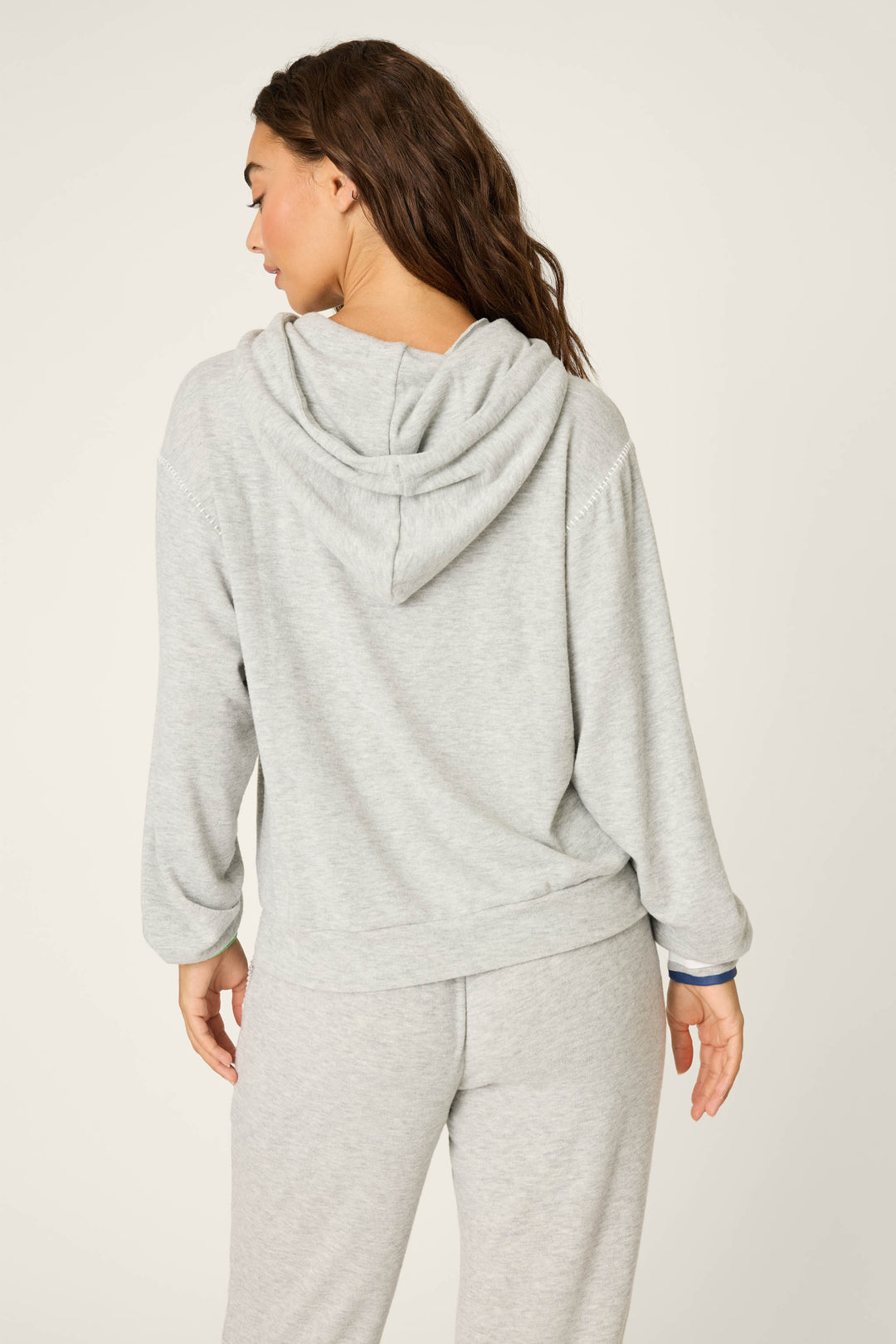 A woman faces away from the camera, wearing the P.J. Salvage C'est La Vie Heather Grey Hoodie with matching sweatpants. Her head is turned slightly left, left arm relaxed by her side, against a plain light background.