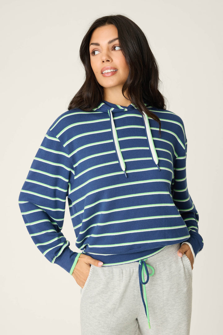 A woman models the P.J. Salvage C'est La Vie Striped Hoodie, featuring blue with green and white stripes, paired with light gray sweatpants. She stands with hands in her pockets against a plain background, gazing slightly to the side.