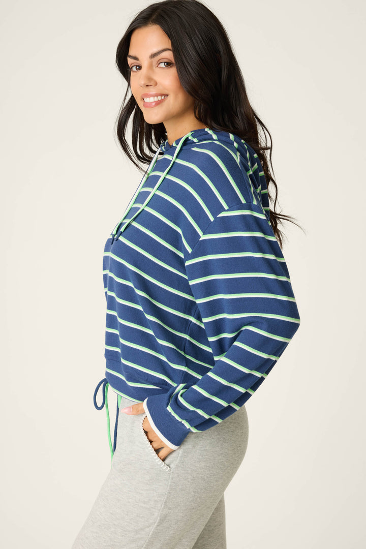 A person with long dark hair smiles sideways with hands in the pockets of a blue and green C'est La Vie Striped Hoodie by P.J. Salvage, paired with light gray pants. The background is plain and light-colored.