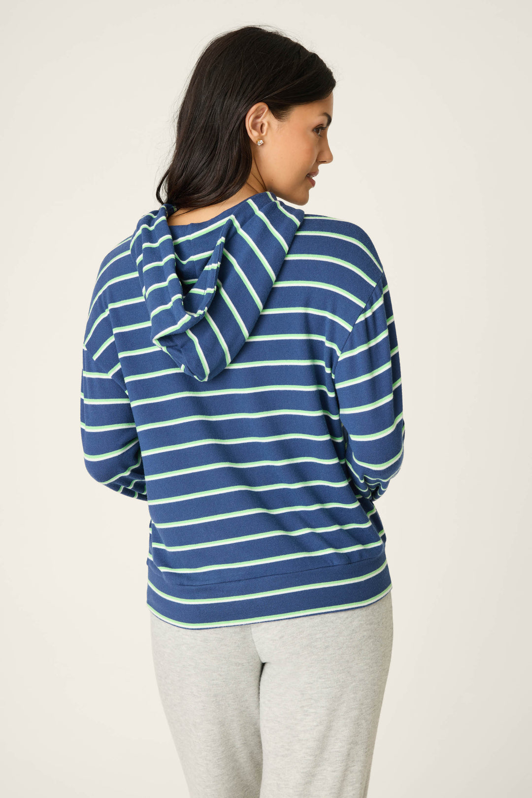 A person with long dark hair is seen from behind wearing the P.J. Salvage C'est La Vie Striped Hoodie in blue with white stripes and light gray pants, standing against a plain off-white background.
