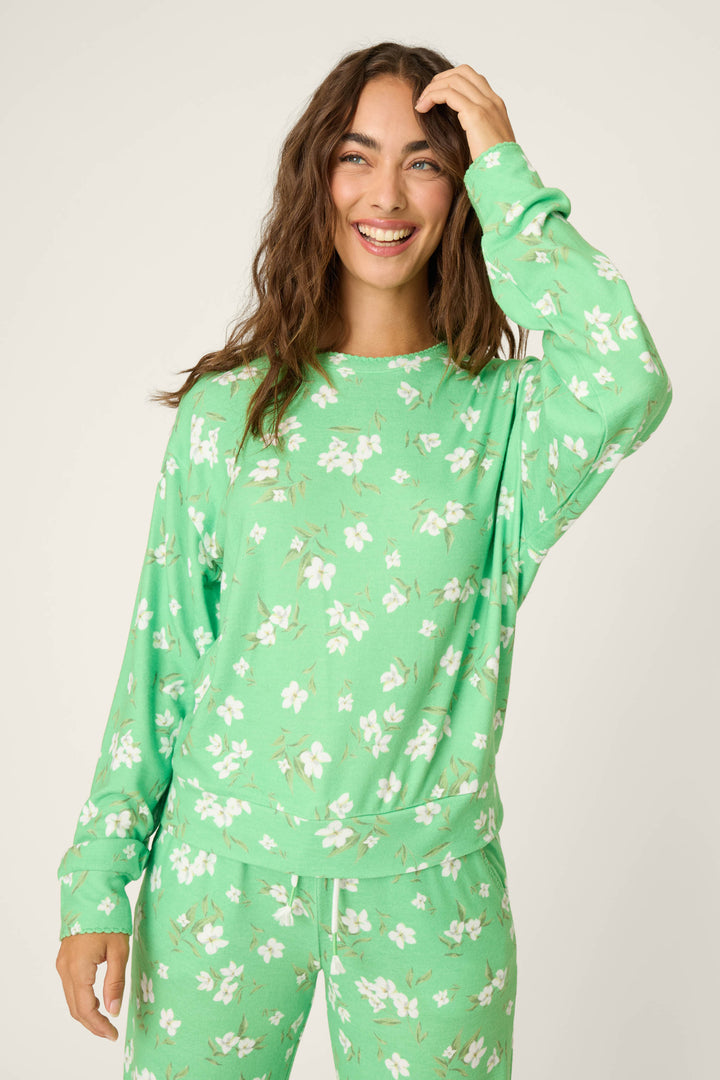 A woman with long brown hair smiles and touches her head while wearing the P.J. Salvage C'est La Vie Floral Print Long Sleeve Top, standing against a plain light background.