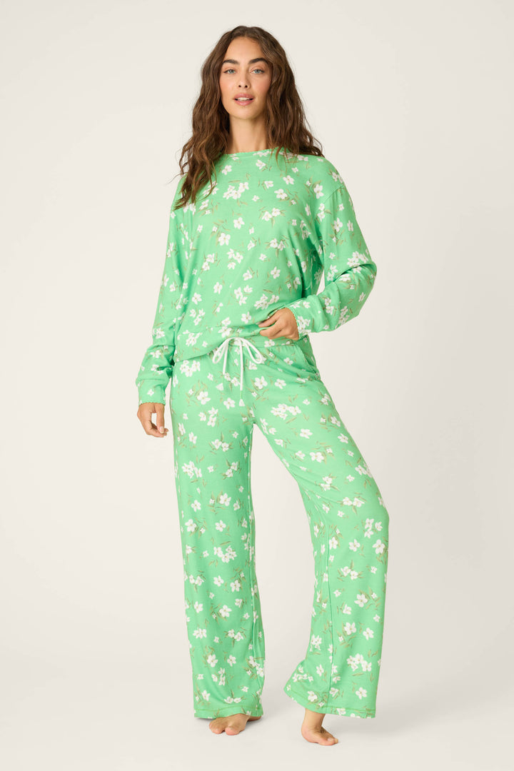 A person stands barefoot in a P.J. Salvage C'est La Vie Floral Print Long Sleeve Top and matching drawstring pants, both light green with a white floral pattern, against a plain off-white background.