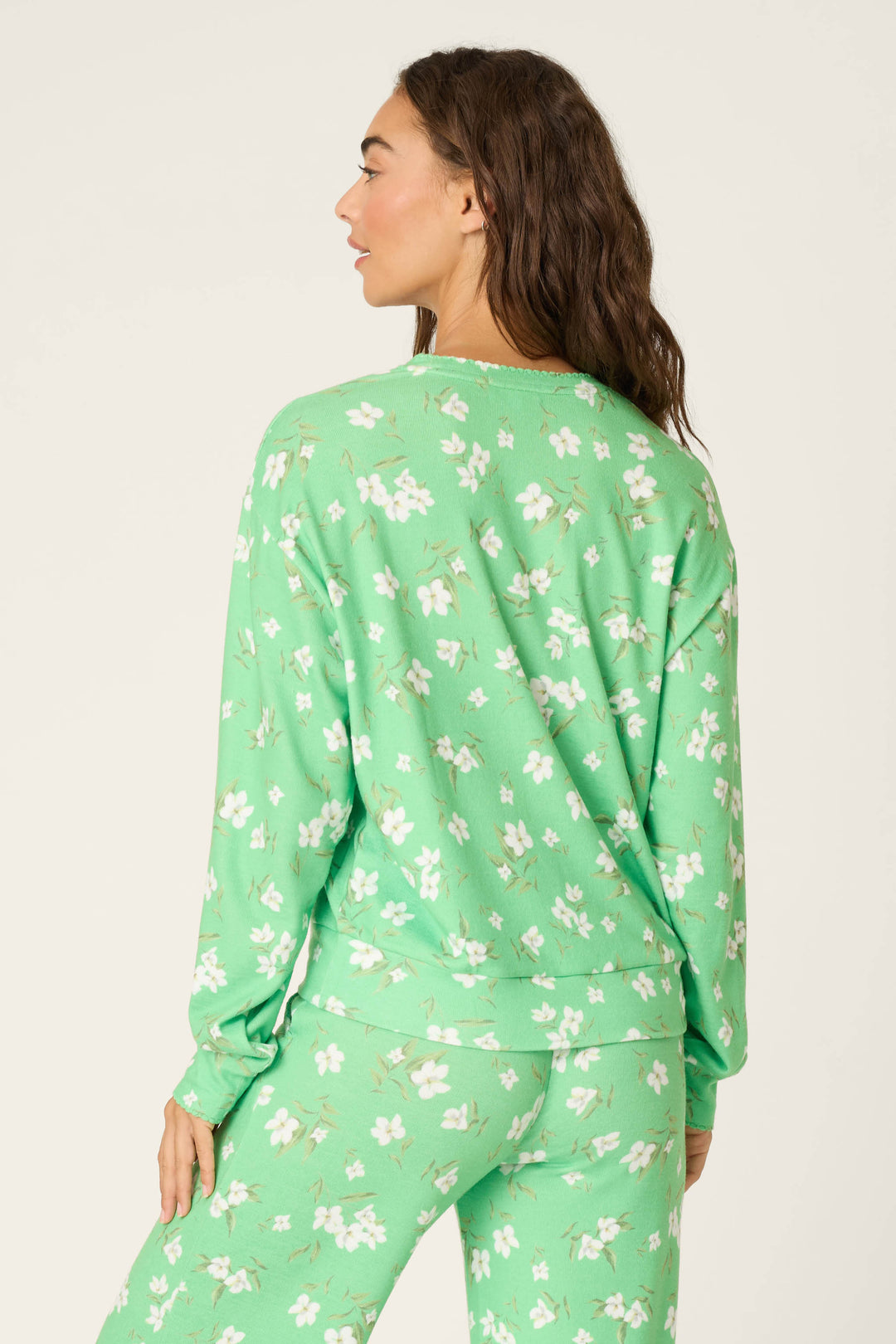 A woman with long brown hair stands facing away, wearing the P.J. Salvage C'est La Vie Floral Print Long Sleeve Top in light green with a white floral pattern, paired with matching bottoms against a plain light background.