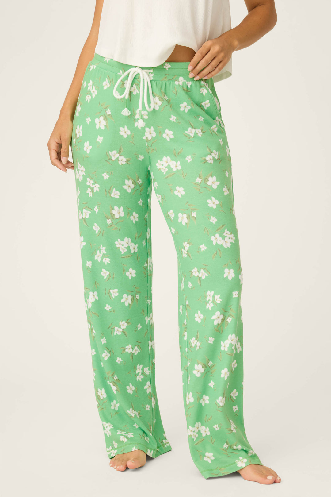 A person stands barefoot wearing P.J. Salvage C'est La Vie Floral Print Open Leg Pants, featuring a green and white floral pattern with a white drawstring, paired with a white cropped top. The image is cropped below the shoulders.